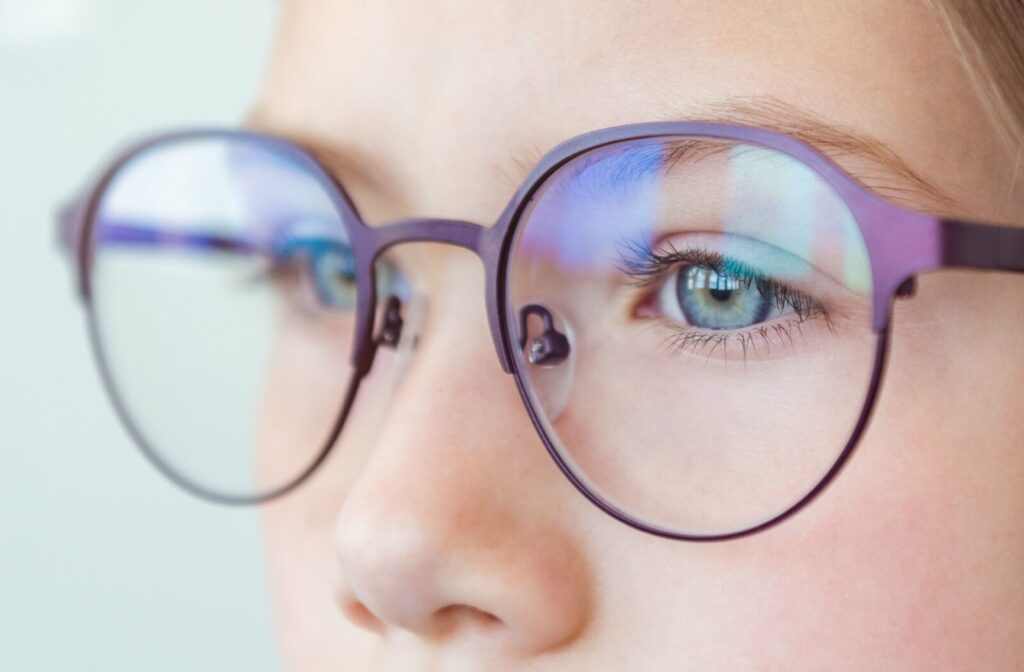 A close-up image of a person wearing eyeglasses in a well-lit room. They have bright blue eyes and a window reflects in the lenses.