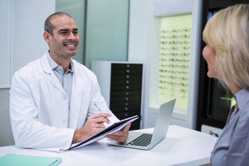 Eye doctor sitting at a desk and smiling while using a notepad and talking to a patient.