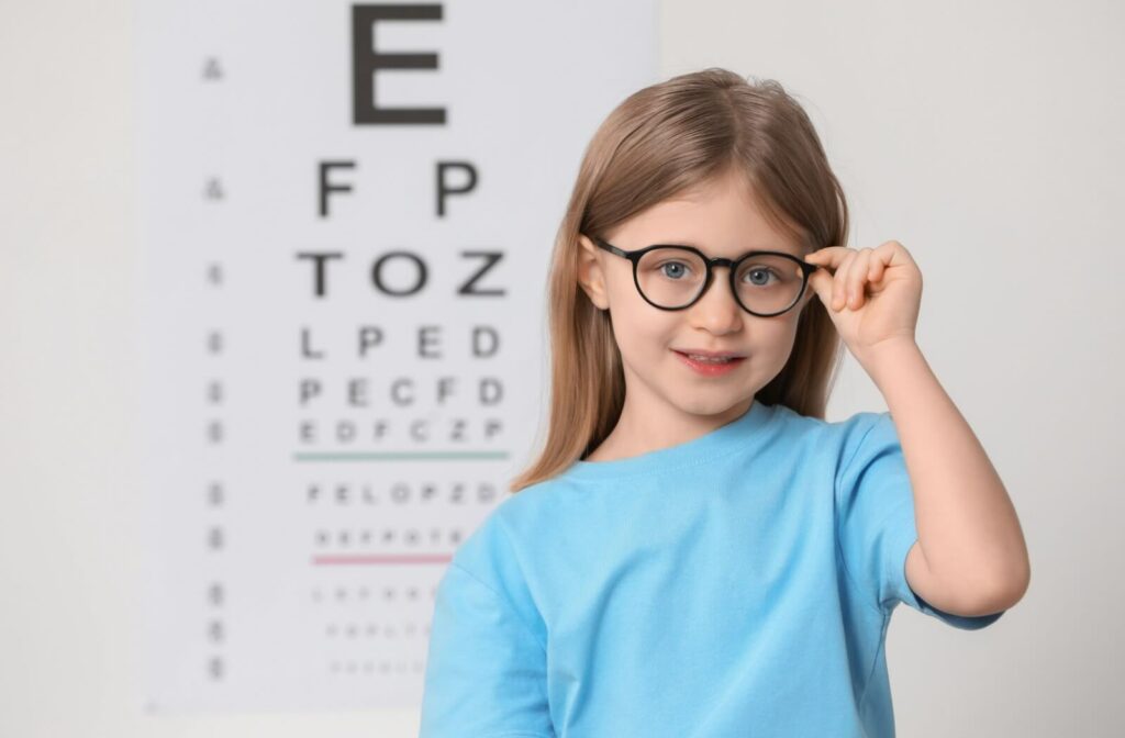 A young child wearing glasses stands in front of a vision chart.