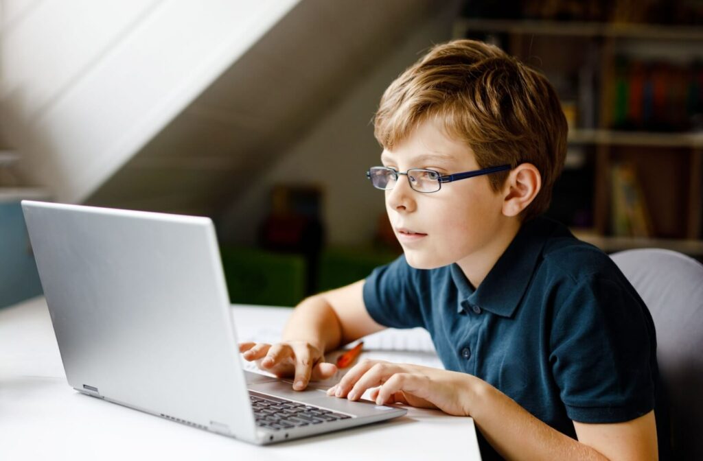 A child wearing glasses works on schoolwork on a laptop.