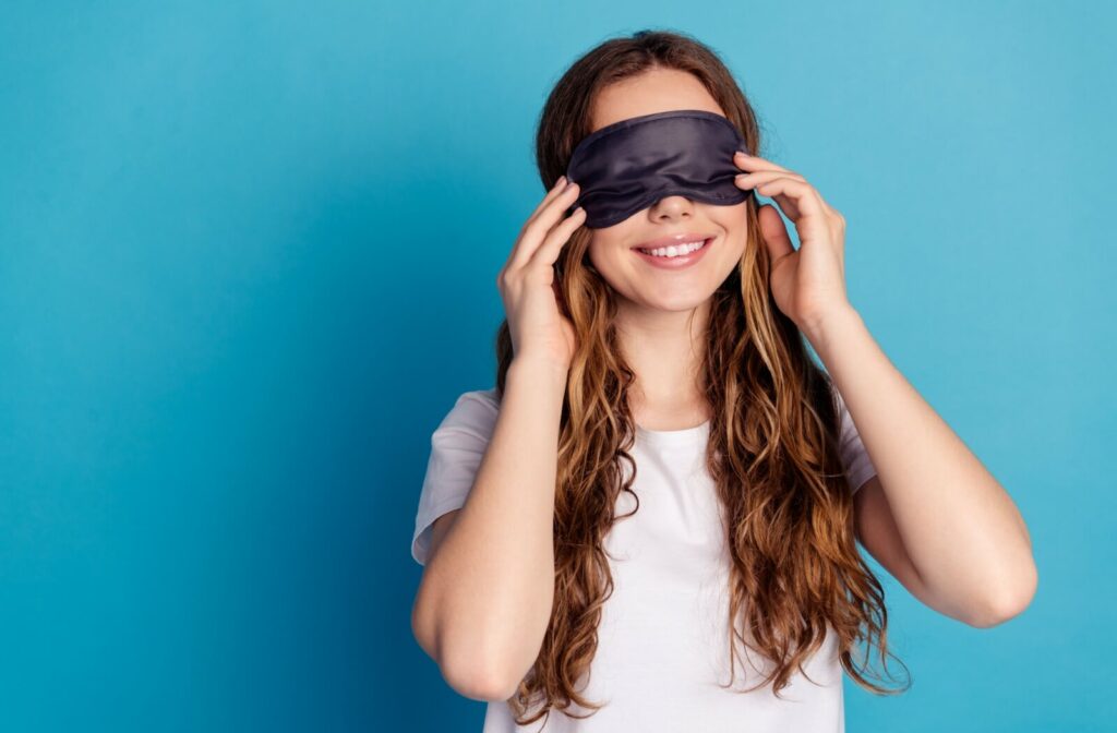 A smiling woman standing in front of a blue background, with her hands on either side of the eye mask she's wearing