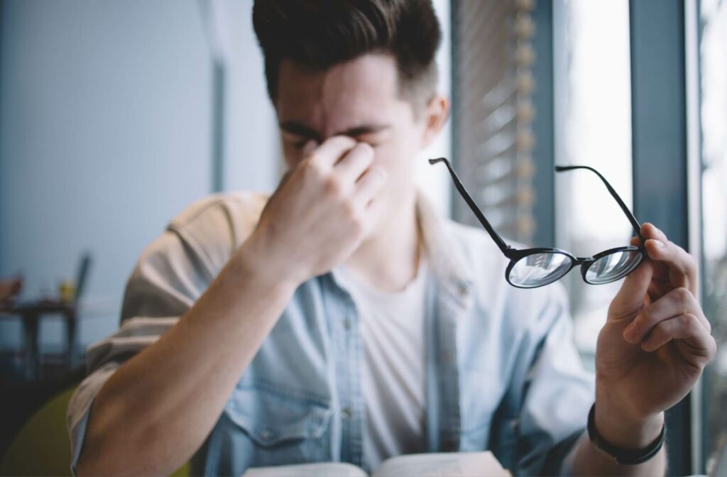 A young adult trying to read a book at their desk, taking off their glasses and rubbing their eyes due to vision problems.